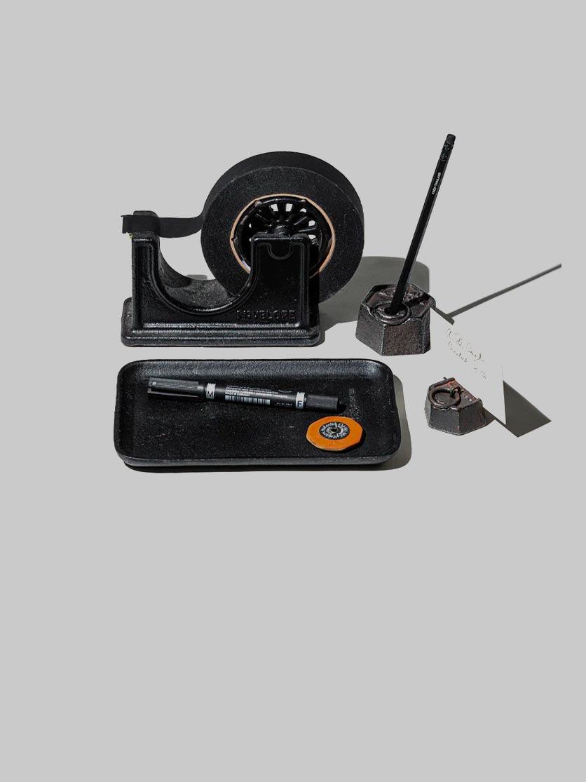 קסת דיו, המכשיר של הסלוטייפ ומגש מתכת בצעב שחור על רקע אפור בהיר בוהק