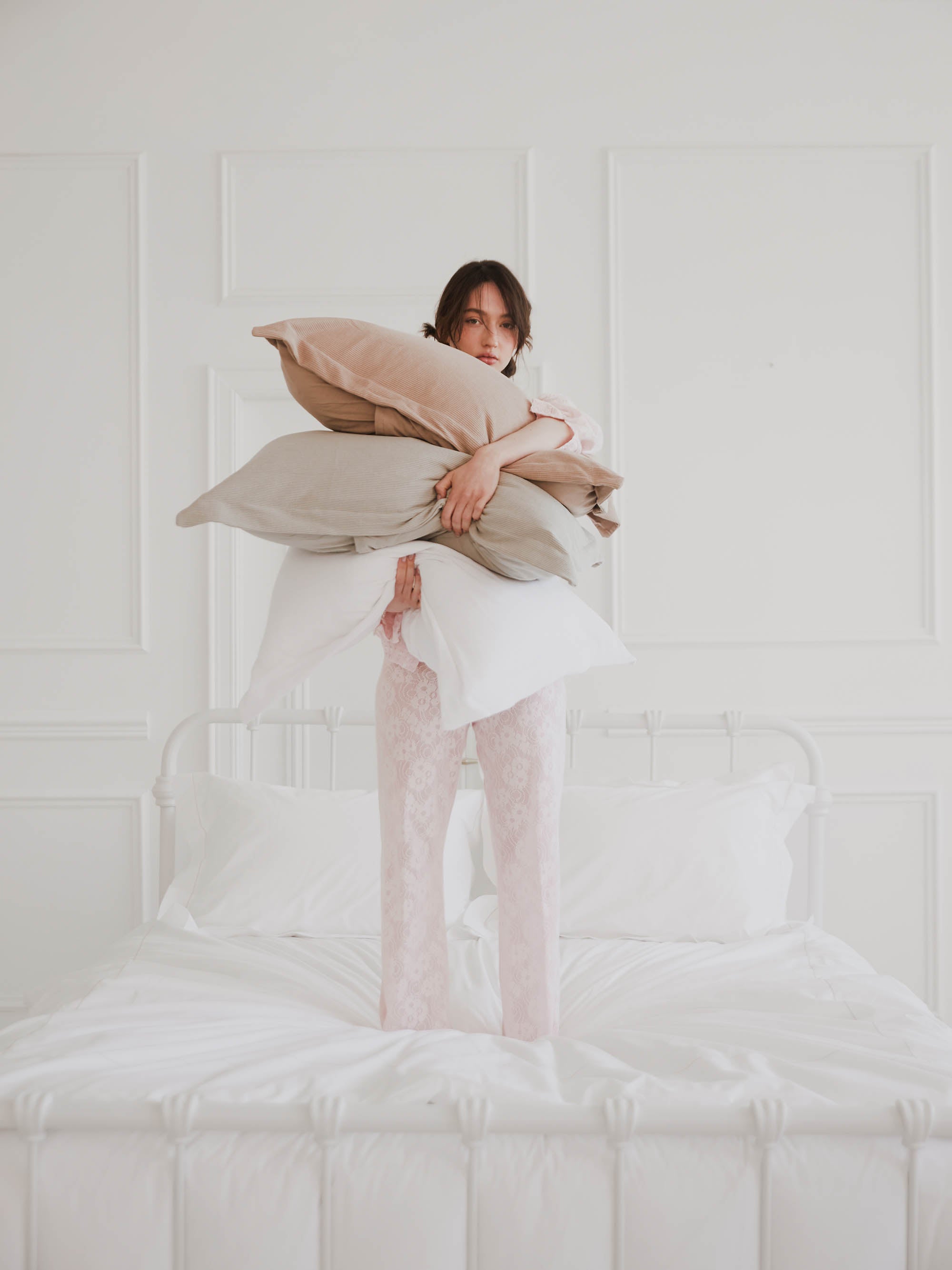 אישה מחזיקה סט כריות עומדת על מיטת ברזל לבנה עם מצעים לבנים 