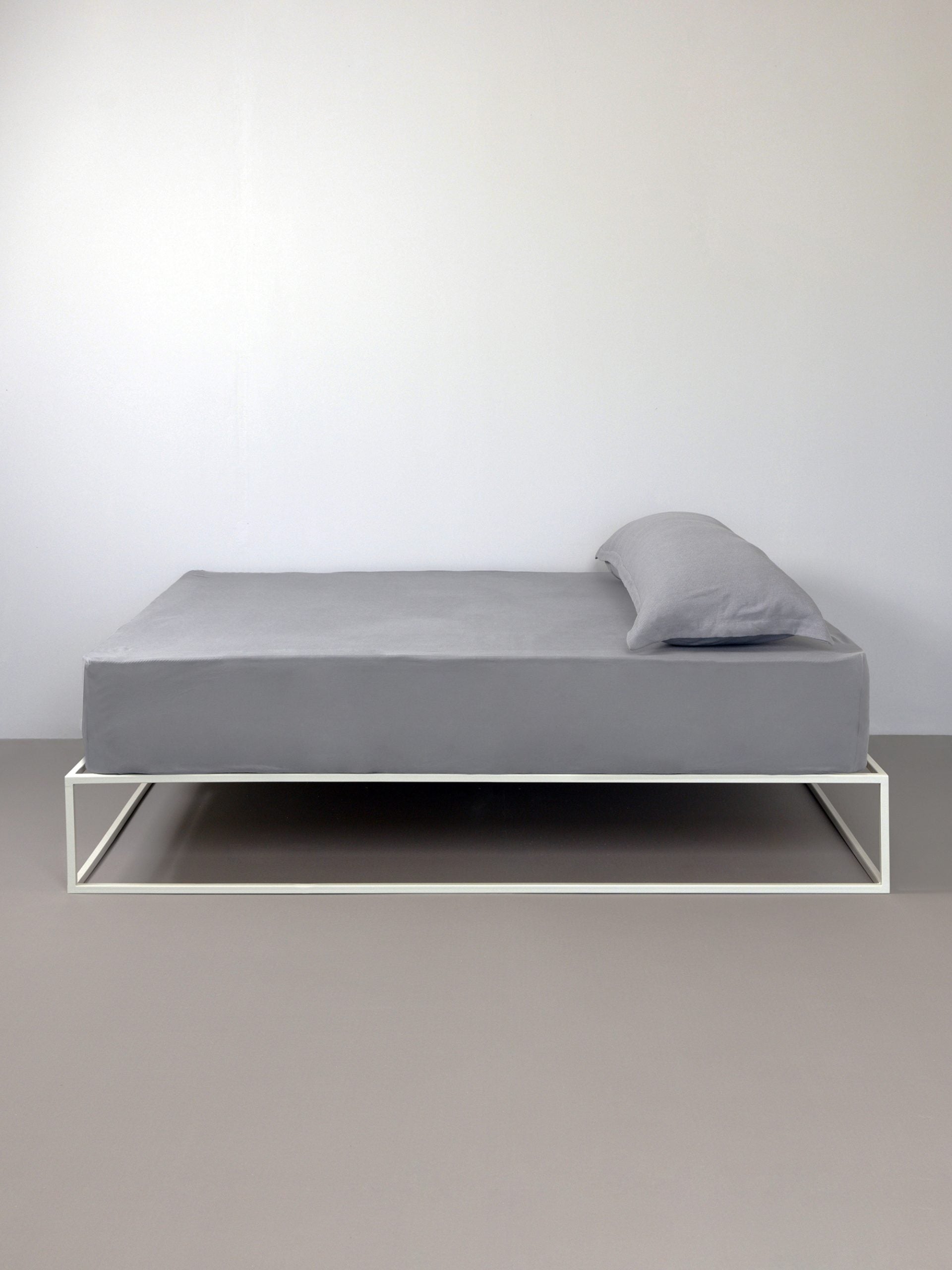 מיטת ברזל לבנה עם סדין, כרית וציפית לכרית ארוכה בצבע אפור בחדר עם קירות אפורים