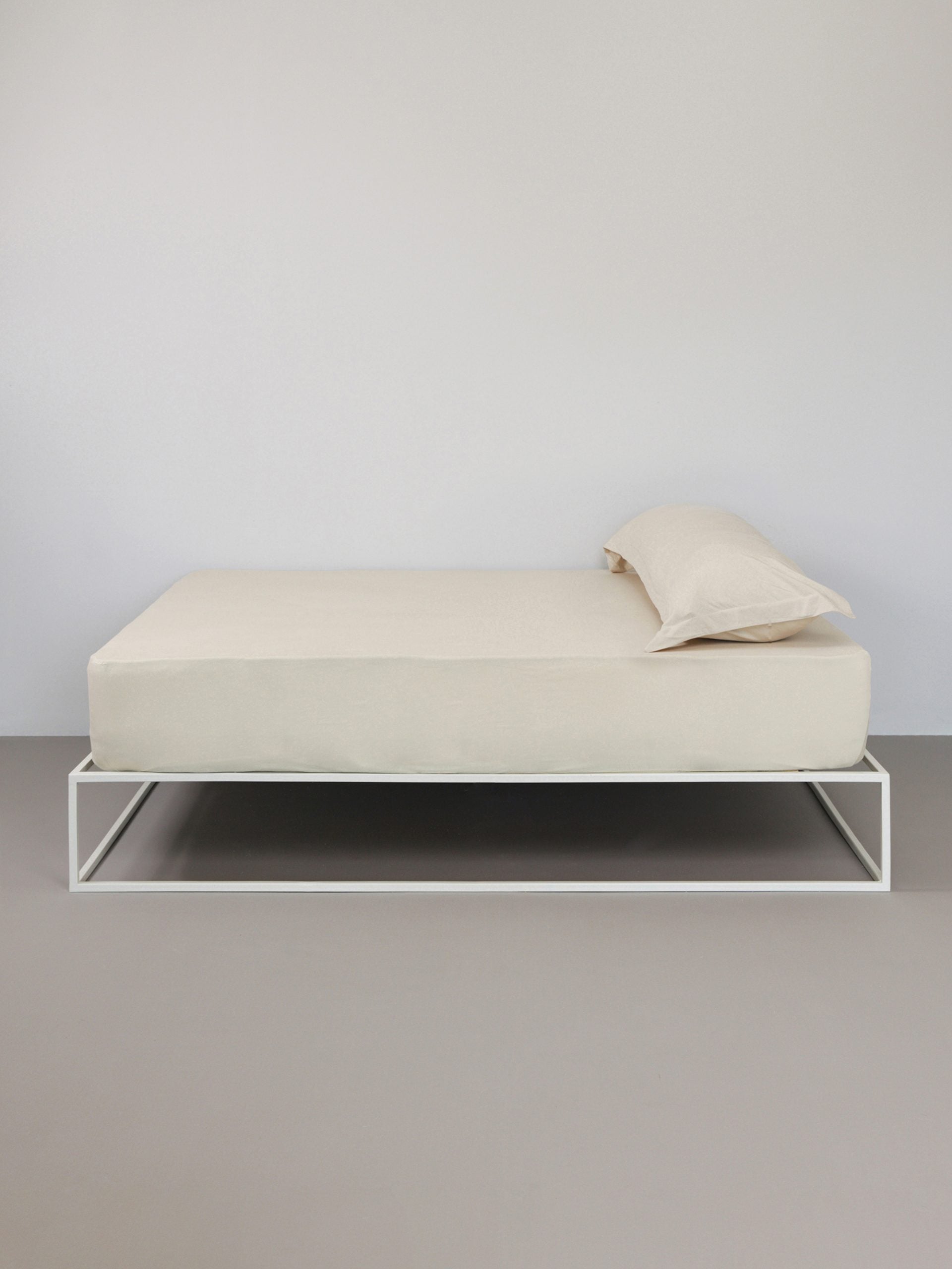 מיטת ברזל לבנה עם סדין, כרית וציפית לכרית ארוכה בצבע אבן בחדר עם קירות אפורים