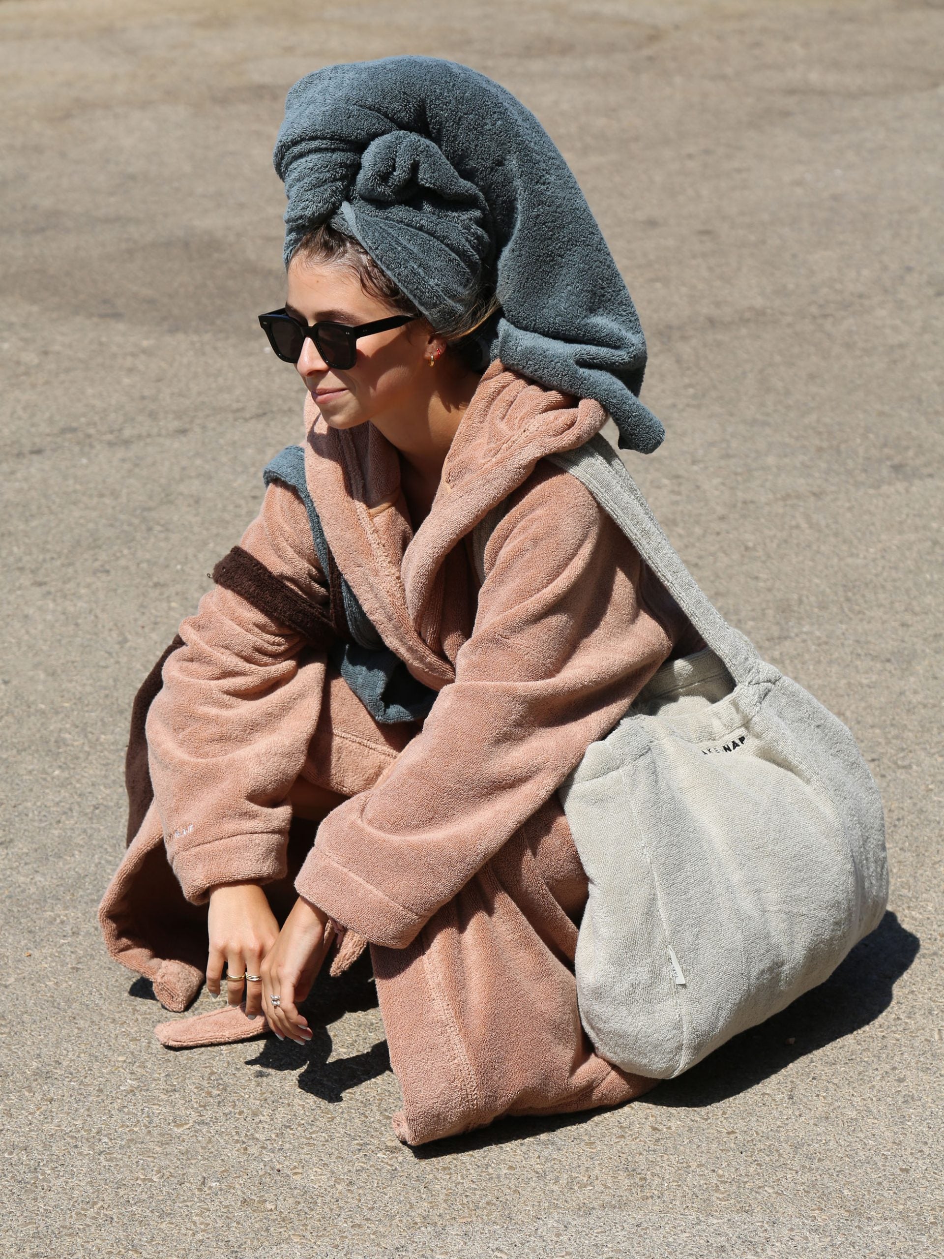 אישה יושבת על משטח אספלט באור יום עם מגבת על ראשה, לובשת חלוק רחצה ועונדת תיק לים