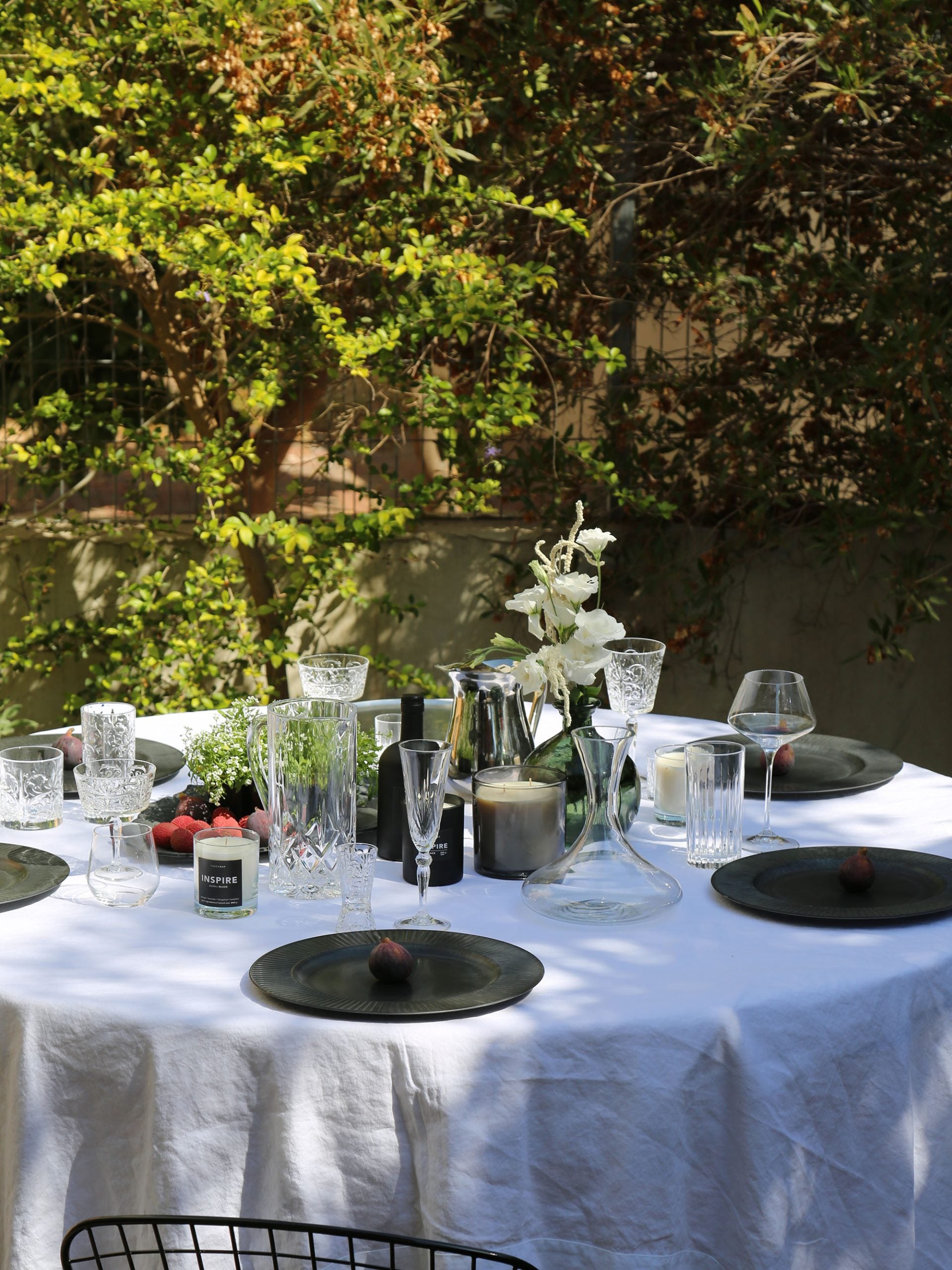 כוסות שמפנייה, מים, נר ריחני, וכד מים מזכוכית על שולחן ערוך עם מפה לבנה וצלחות בחצר באור יום