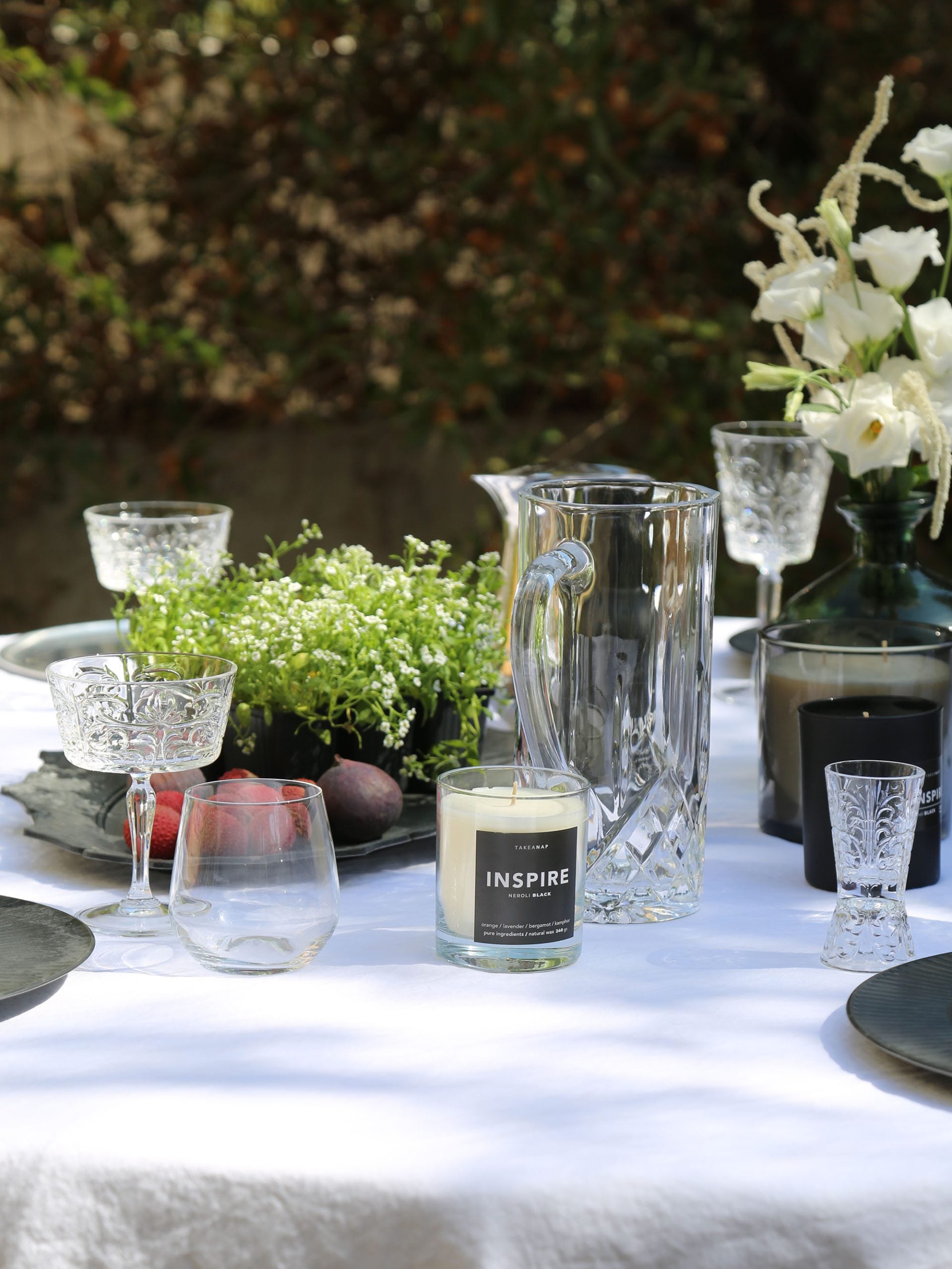כוסות שמפניה, מים, נר ריחני, וכד מים מזכוכית על שולחן ערוך עם מפה לבנה וצלחות בחצר באור יום