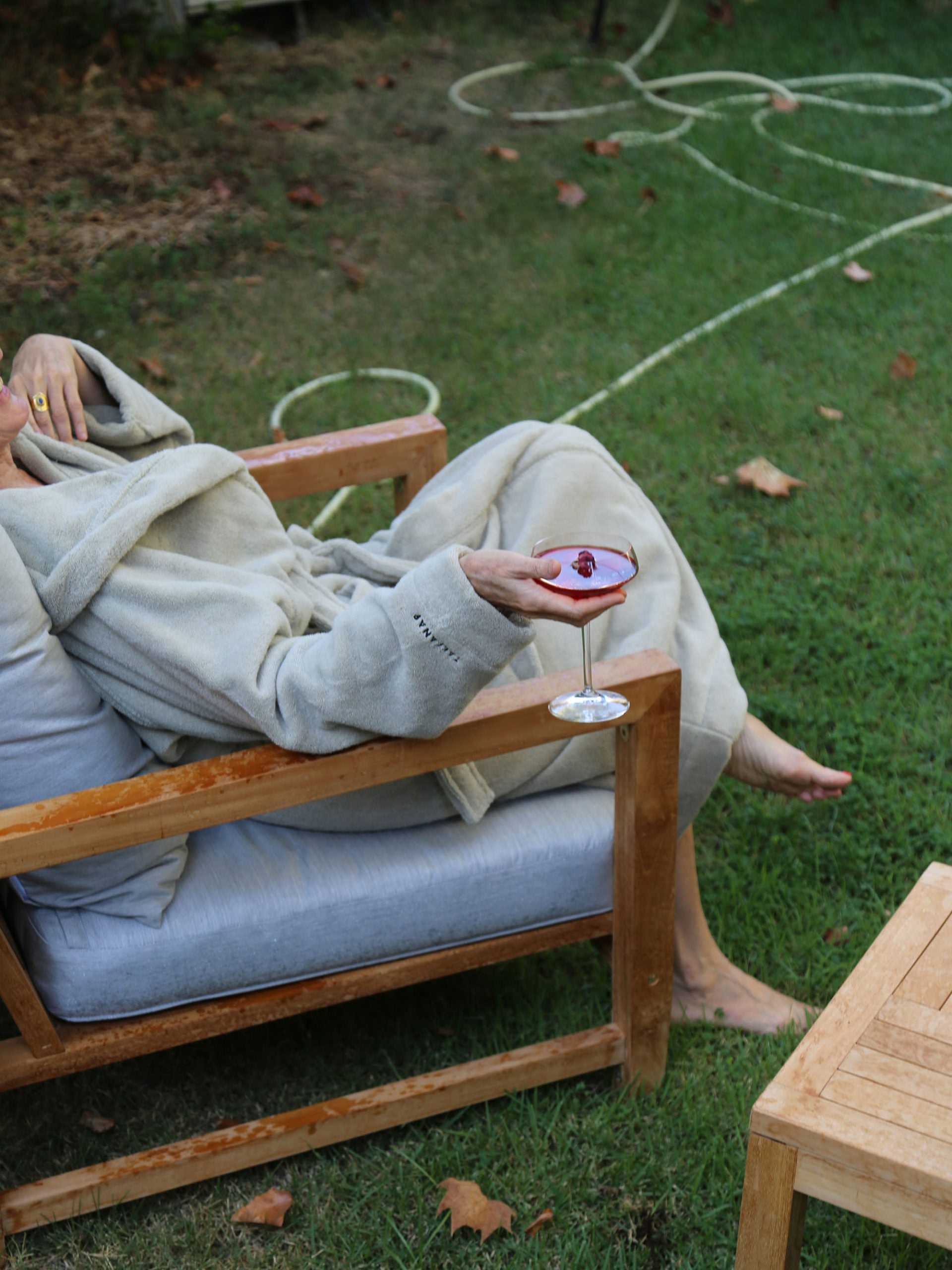  אישה שלובשת חלוק רחצה יושבת על כיסא עץ בגינה שלה ומחזיקה בידה כוס יין עם יין אדום