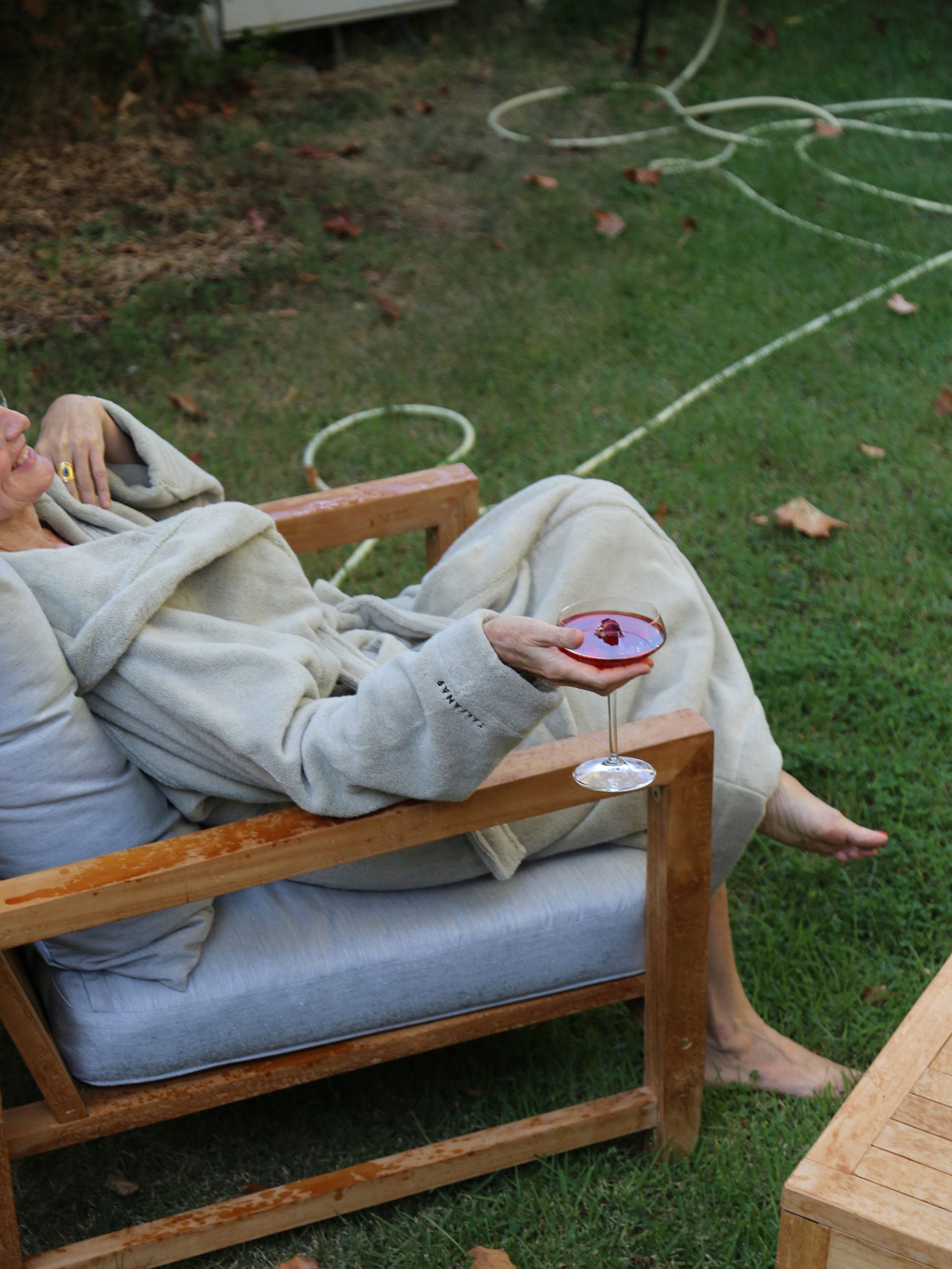 אישה יושבת על כיסא בחצר עם חלוק רחצה וכוס שמפנייה בידה