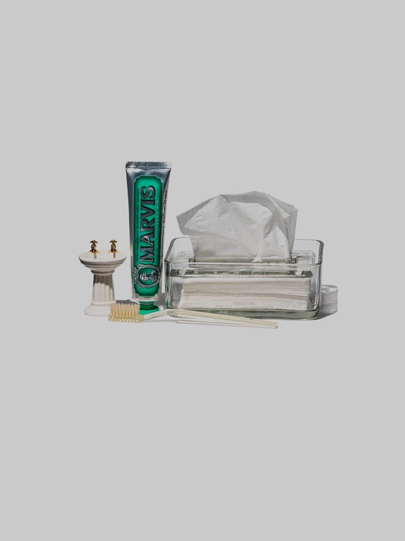 קופסת טישו מזכוכית עם נייר טישו, משחה ומברשת שיניים על רקע אפור