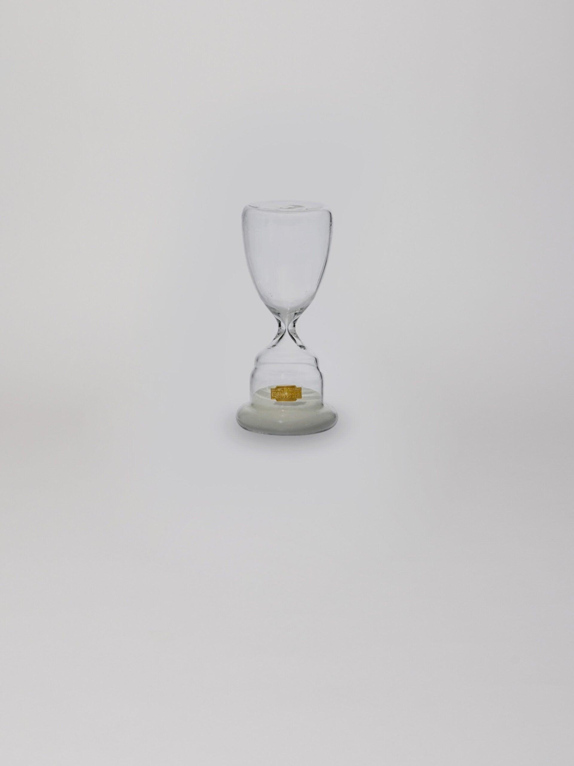 שעון חול מזכוכית בצבע לבן עם חול בחלקו התחתון על עומד על רקע אפור בהיר