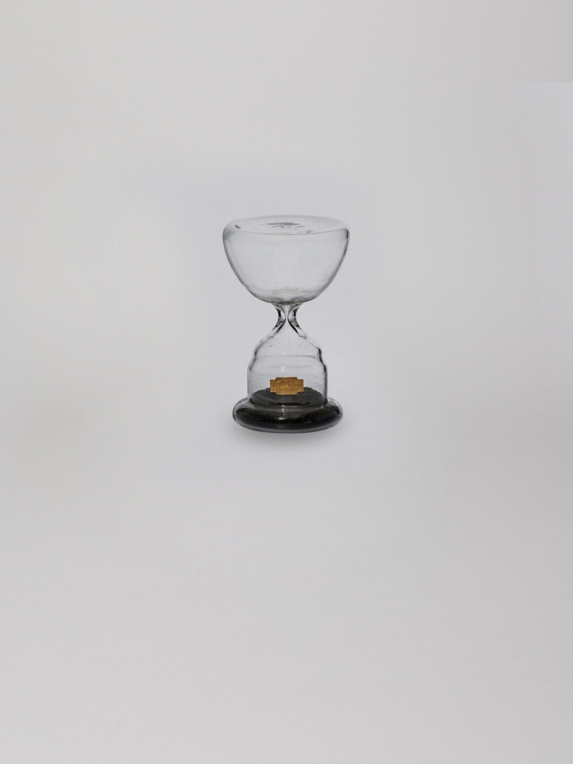 שעון חול מזכוכית בצבע שחור עם חול בחלקו התחתון על עומד על רקע אפור בהיר