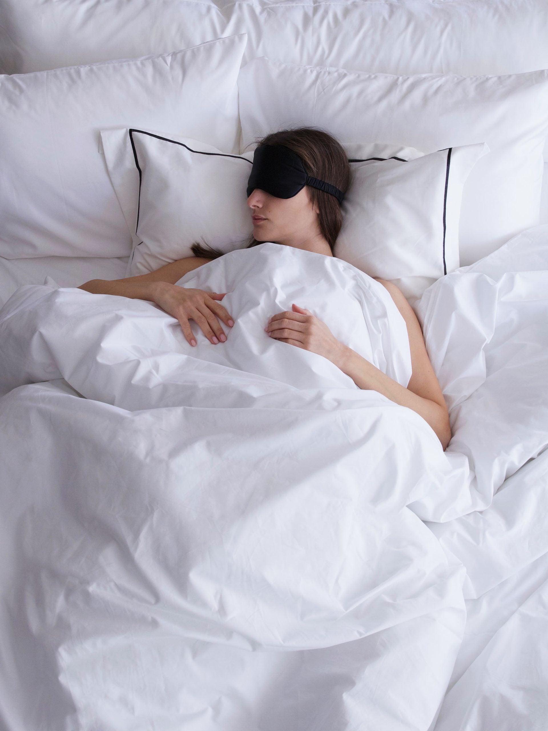 אישה עם כיסוי עיניים שכובה במיטה עם סט כריות לבנות עם ציפיות מכוסה בשמיכת פוך לבנה