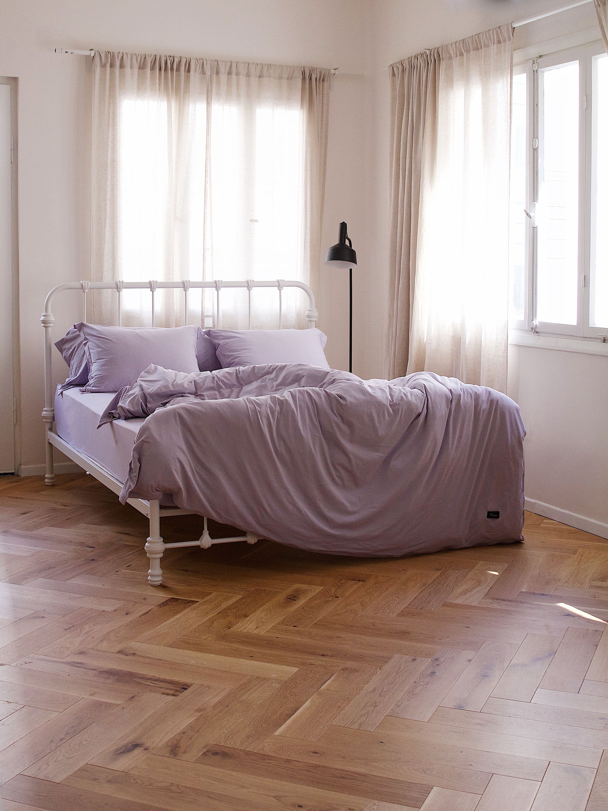 מיטת ברזל לבנה עם מצעים איכותיים בחדר עם פרקט חלונות לבנים והרבה שמש