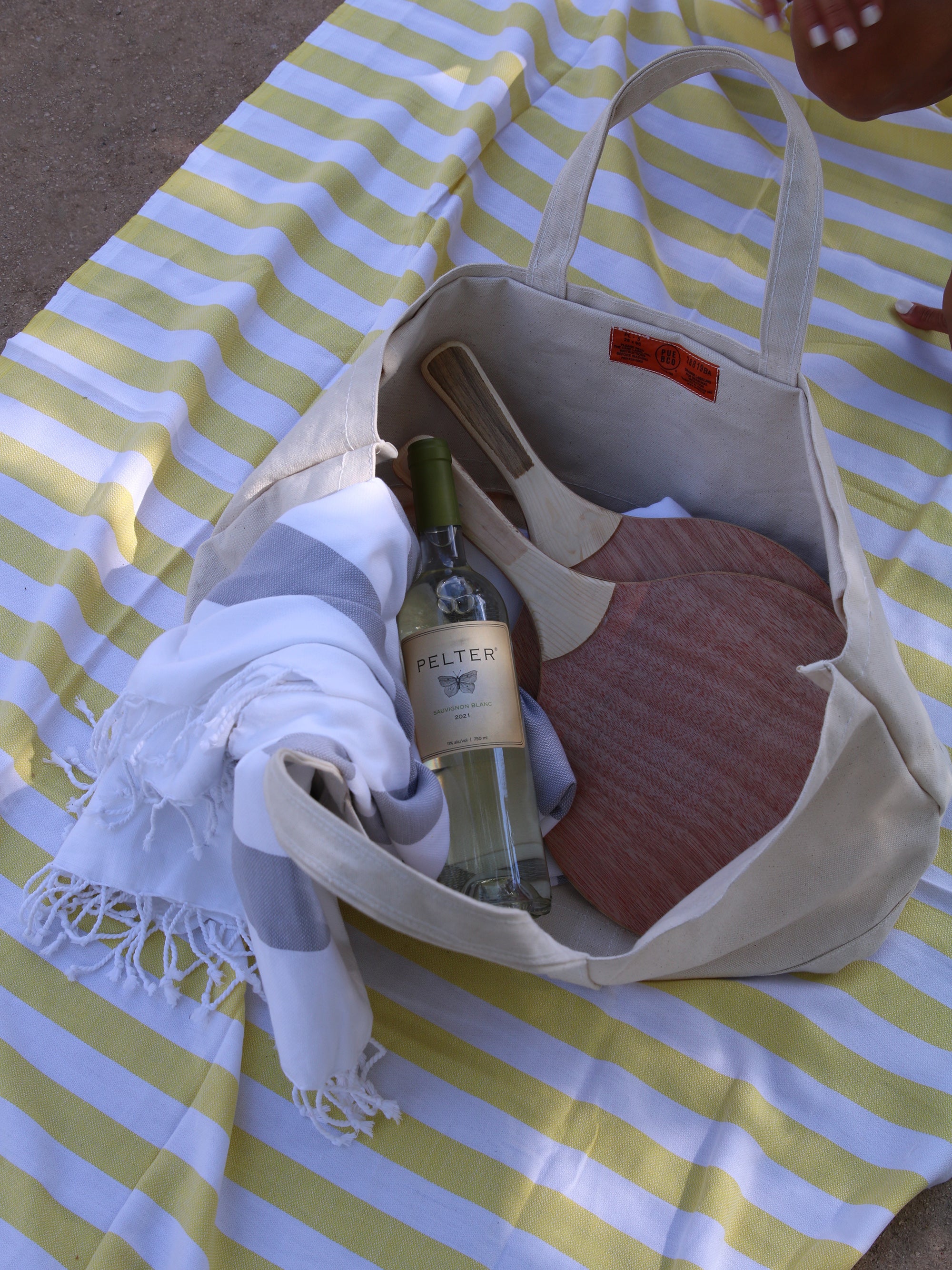 תיק קנבס פתוח שבתוכו מטקות עץ, בקבוק יין לבן ושמיכת חוף לבנה מונח על פשטמל צהוב 