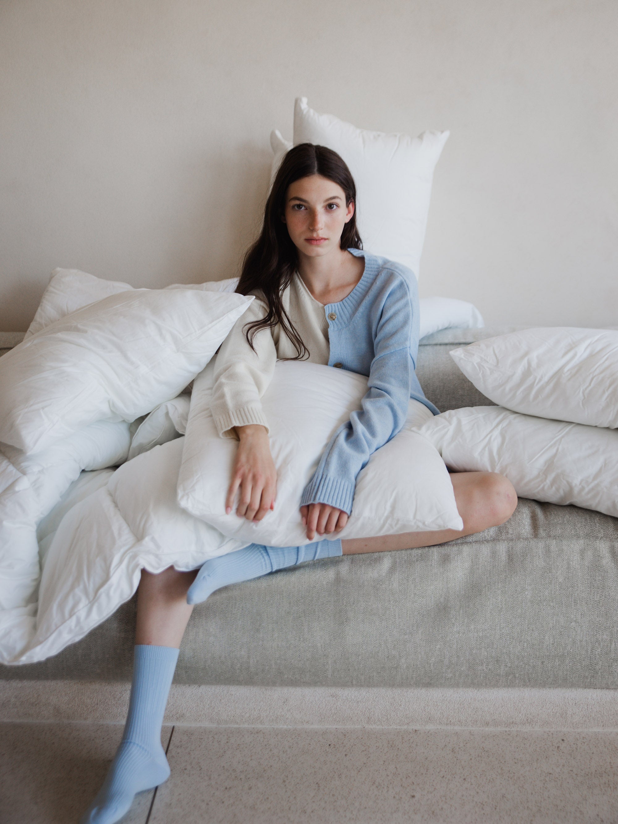 אישה על ספה מחזיקה כרית שינה שמסביבה מונחות עוד כריות למיטה 