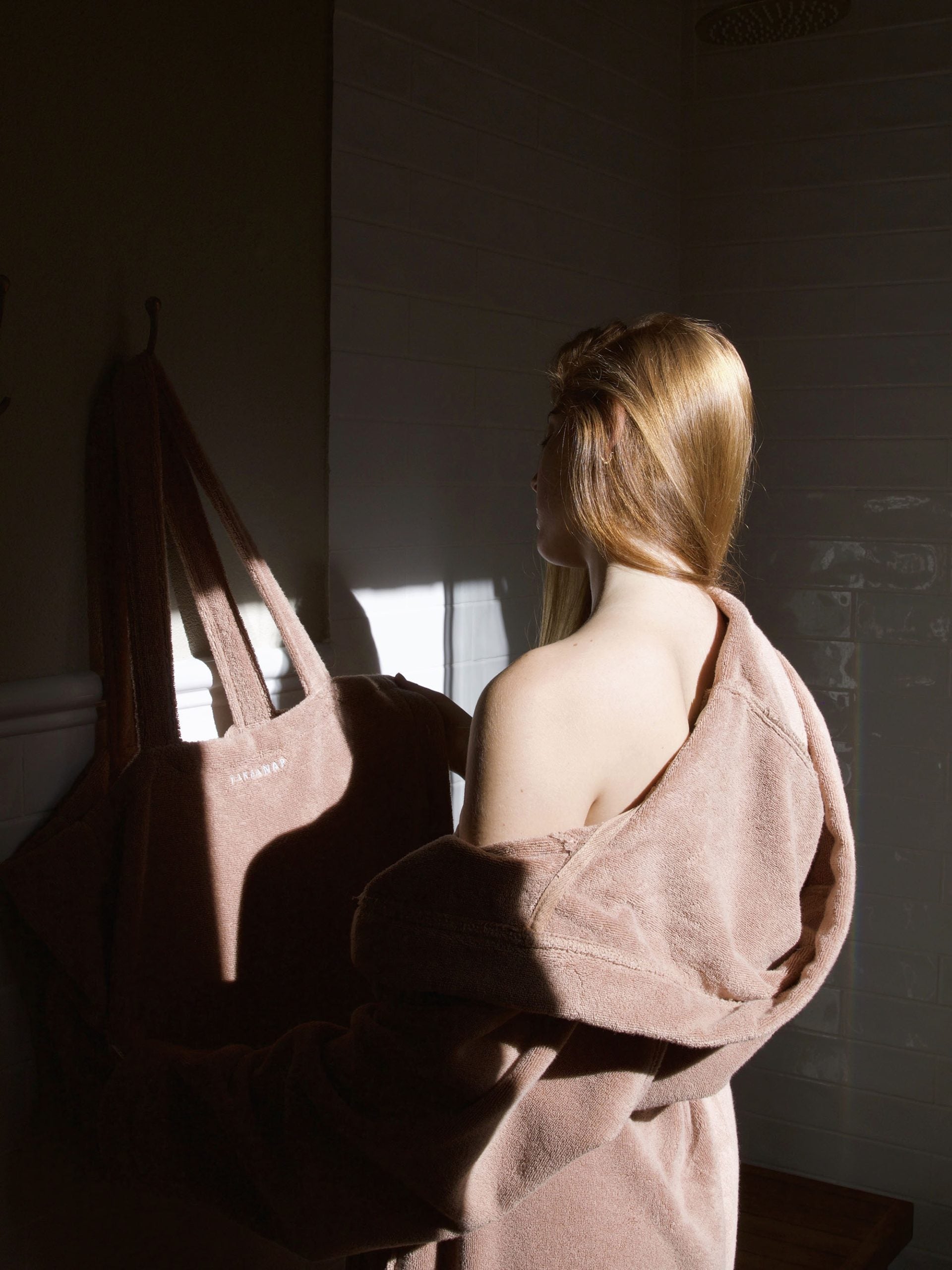 אישה שלובשת חלוק רחצה בצבע לאטה מסתכלת ונוגעת בתיק יםבתיק ים שתלוי על הקיר