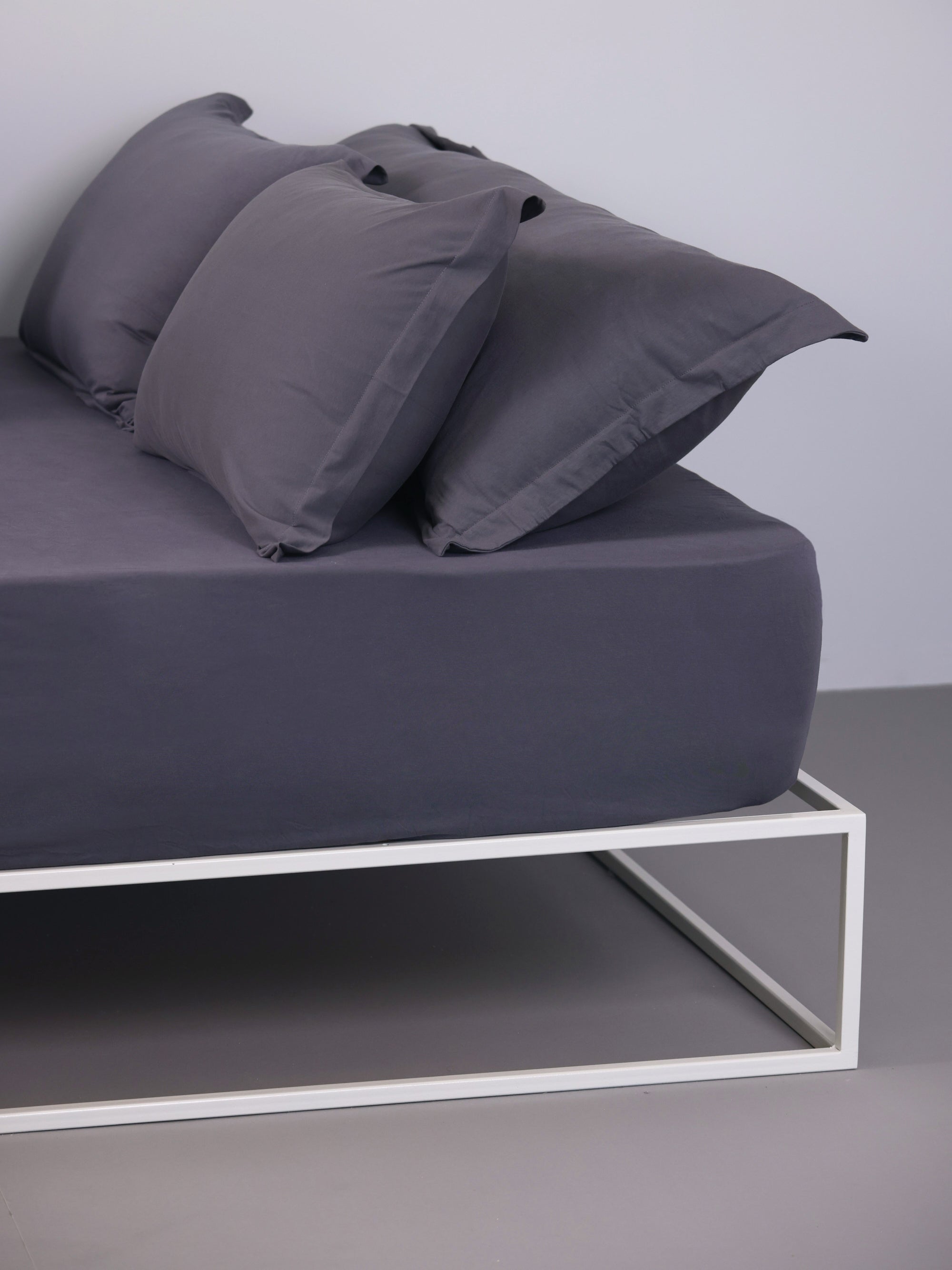 מיטת ברזל לבנה עם סדין, כרית וציפית לכרית ארוכה בצבע גרפיט בחדר עם קירות אפורים