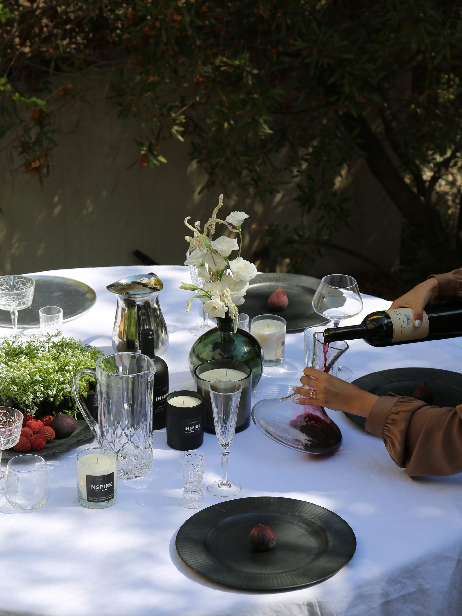 אישה מוזגת יין לתוך דקנטר יין מזכוכית על שולחן ערוך עם מפה לבנה שעליו כוסות שמפניה, צלחות מעוצבות ונרות ריחניים