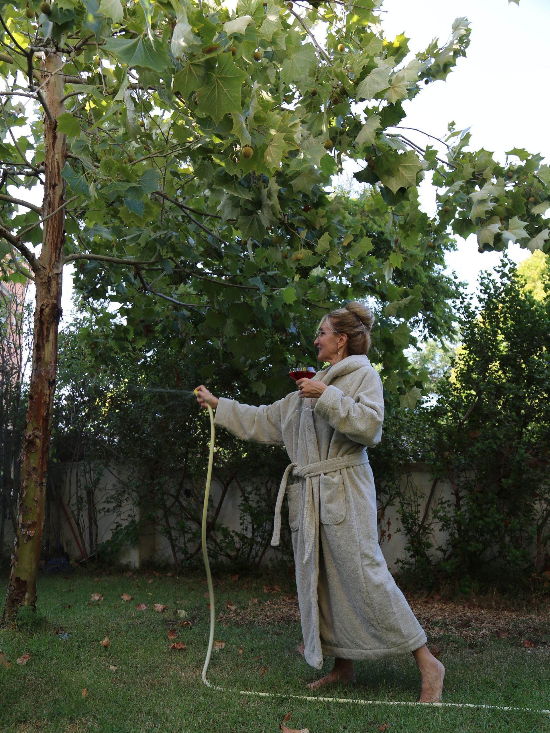 אישה שלובשת חלוק רחצה נמצאת בגינה ומשקה עם צינור עץ 