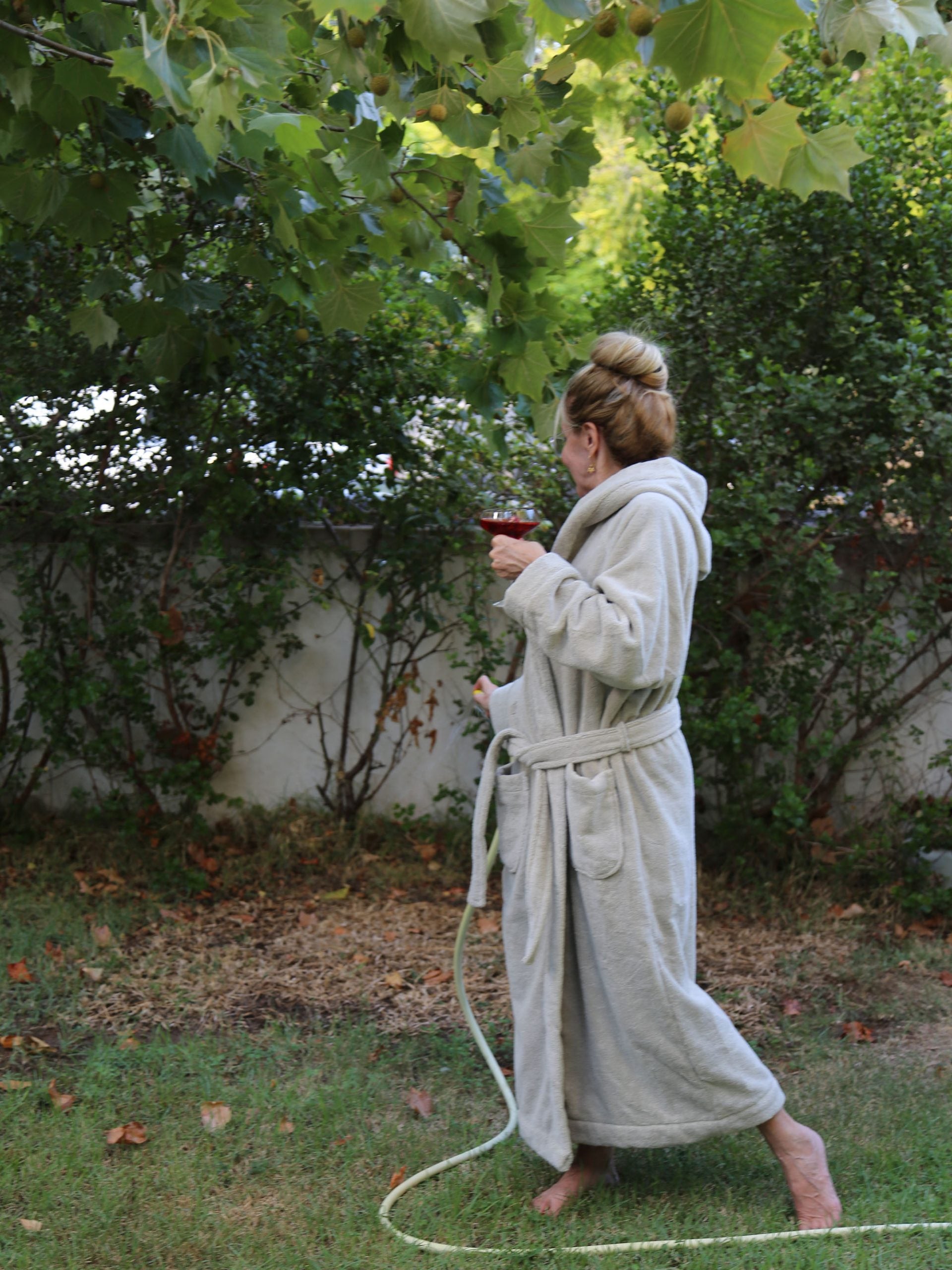 אישה לובשת חלוק רחצה כשבידה כוס שמפנייה מתחת לעץ בחצר בית 