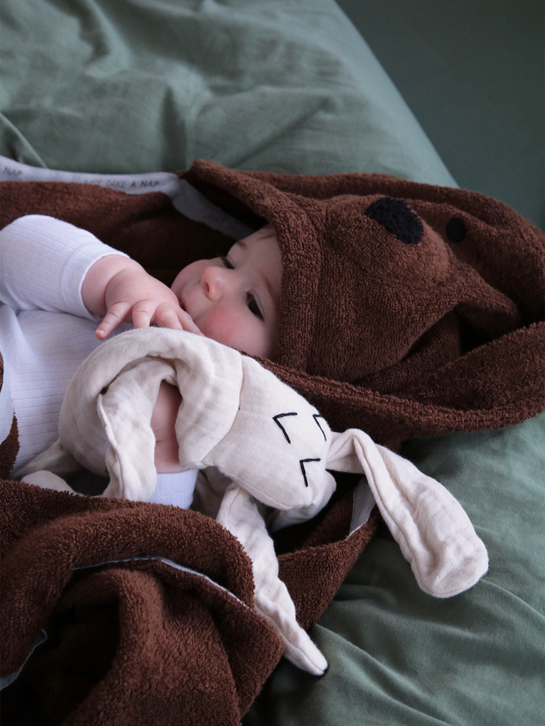 תינוק שמחזיק חפץ מעבר לבן  על מיטה עם מצעים מכוסה במגבת חיבוקי בצבע חום