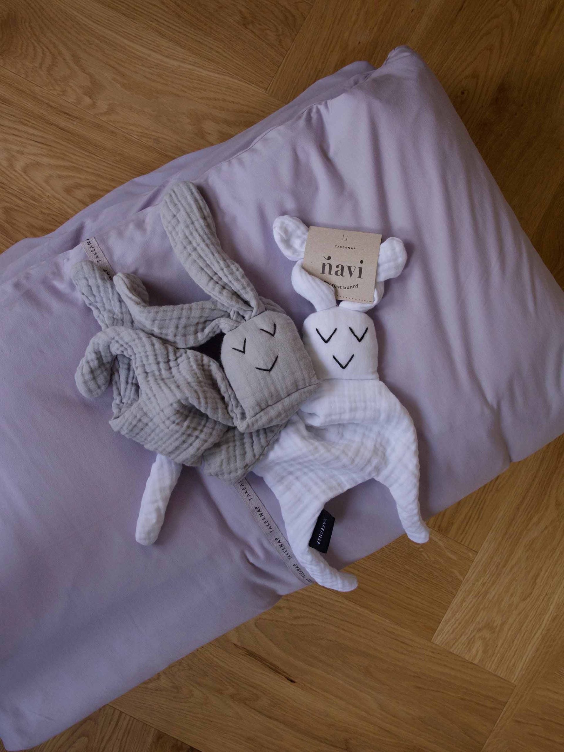 חפץ מעבר נבי ארנבי Dream לבן ואושן מונחים על כרית למיטה שהיא מונחת על רצפת פרקט