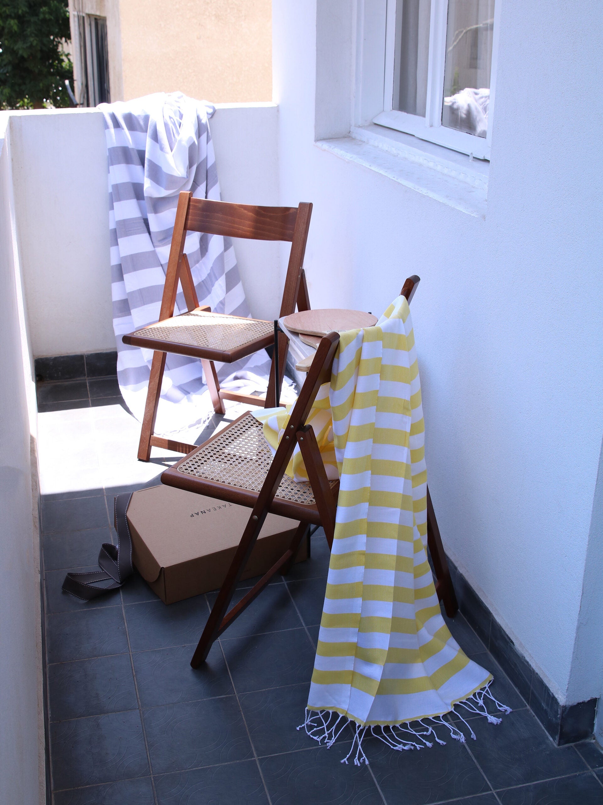 מרפסת בית עם שני כיסאות עץ שעל קיר המרפסת מונחת שמיכת חוף אפורה ועל כיסא מונחת שמיכת חוף צהובה
