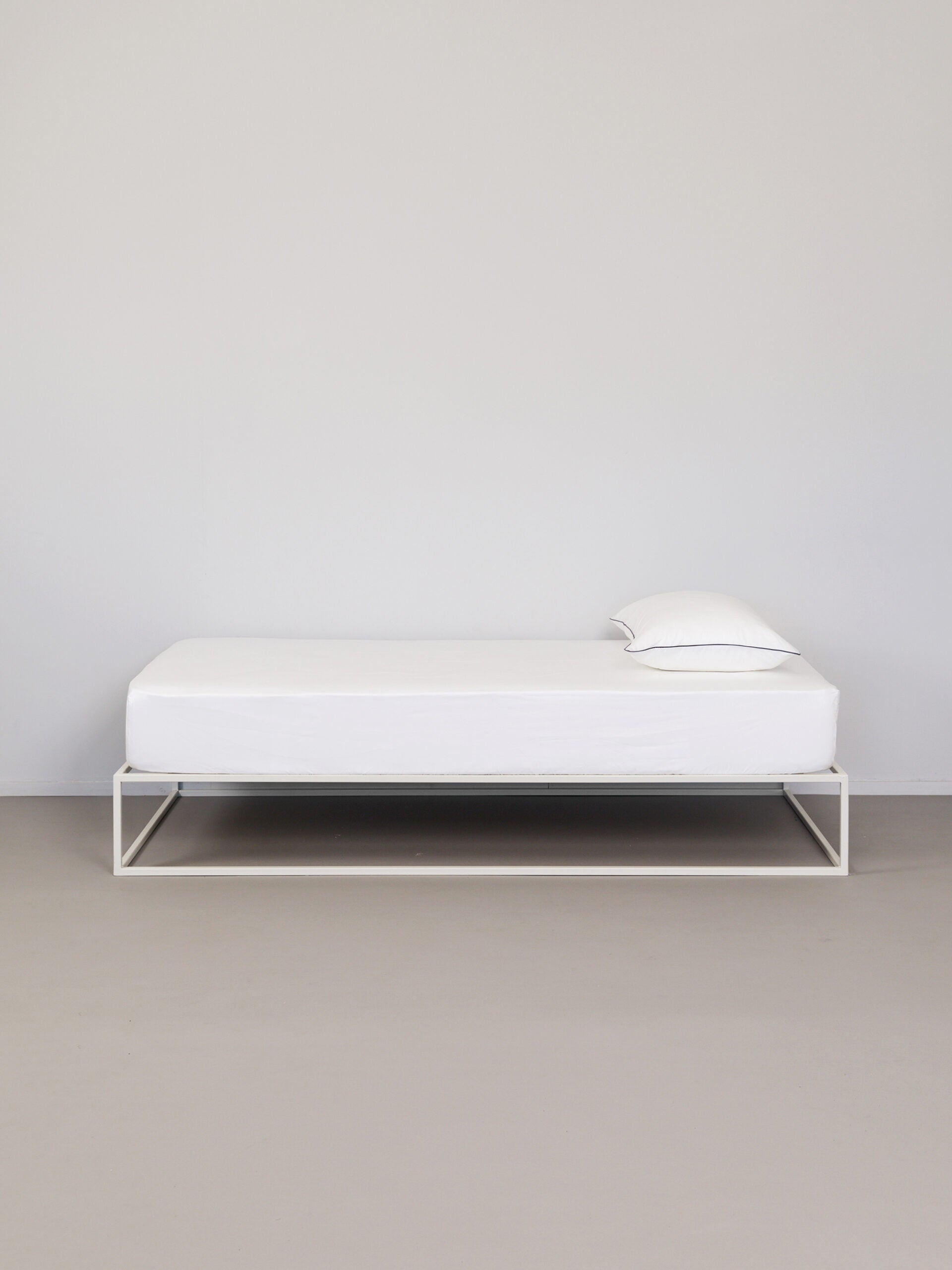 מיטת ברזל לבנה עם סדין לבן וכרית עם ציפית לכרית לבנה עם פס שחור בצדדיו בחדר אפור בהיר בוהק